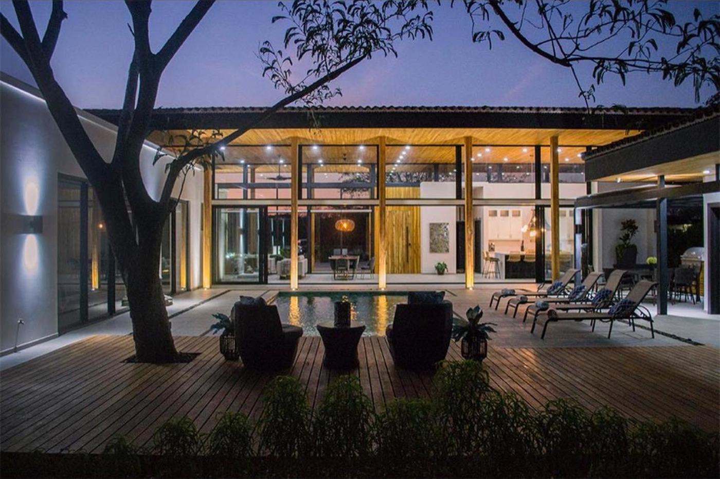 Glass-fronted home at dusk with warm lighting, a timber deck, and outdoor seating